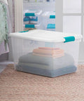 Super Tight Fit 64 Quart Clear Plastic Storage Bins 72 Pack With Snappy Hinged Lids stacked with folded blankets in a bedroom.