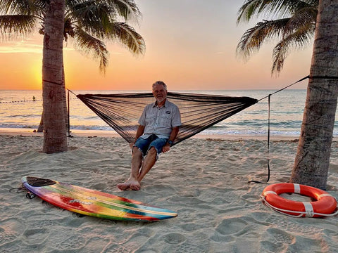 Ultralight Pocket Hammock For Solo Adventures set up between two palm trees on a beach at sunset, supports up to 330lbs.