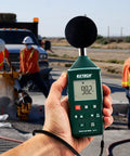 Compact Digital Sound Level Meter With Plus Minus 1dB Accuracy, Class 2, Backlit LCD used on job site with workers in background.