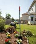 Dual Hoe And Rake Garden Tool With 58 Inch Fiberglass Shaft standing in a garden bed with flowers and mulch next to a house