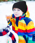 Child in a colorful jacket staring at camera, holding a carrot while wearing a black knit hat in snow. No balaclava depicted.