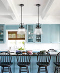 Black Farmhouse Pendant Light With Clear Glass Schoolhouse Style hanging over a kitchen island with blue cabinets and black chairs.