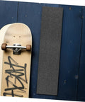 Two Pack Black Skateboard Grip Tape Sheets 33 Inch By 9 Inch Anti Skid Sandpaper next to a skateboard on a wooden surface