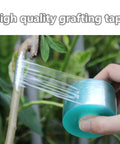 Hand using grafting tape from Grafting Tool Kit For Garden Pruning And Budding Dope Replacement Blades Pro Grade Plant Branch Vine And Fruit Tree Cutting Tools Scissors For Citrus Apple Peach Persimmon And Other Fruit Or Flower Trees High Survival