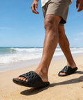 A person walking on a beach wearing Drainage Holes Shower Slippers With Quick Drying Sole For Men And Women Open Toe Bath Shoes Soft Cushioned Footbed Ultra Comfy For Home Gym Pool And Spa Black 44.45.