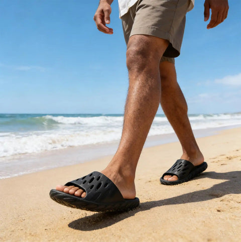A person walking on a beach wearing Drainage Holes Shower Slippers With Quick Drying Sole For Men And Women Open Toe Bath Shoes Soft Cushioned Footbed Ultra Comfy For Home Gym Pool And Spa Black 44.45.