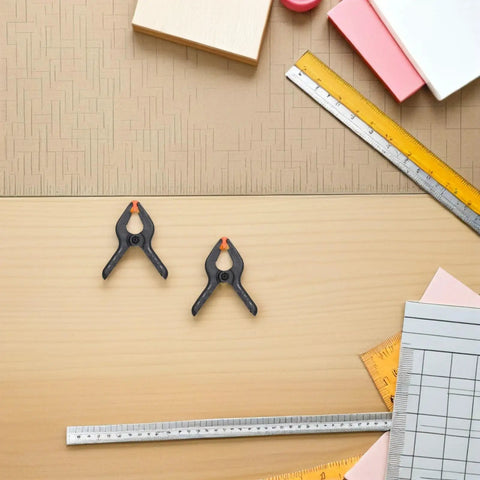 Top Tier Heavy Duty Spring Clamps Set For Woodworking And Photography Backdrops, Elastic Plastic Clips With Compact Design Great For Any Creative Project. Two black spring clamps on a wood surface with rulers and paper samples