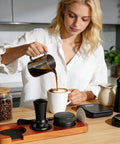Woman pouring coffee using Portable Espresso Tamper Kit For 51Mm Filter Cups on a wooden tray with black accessories.