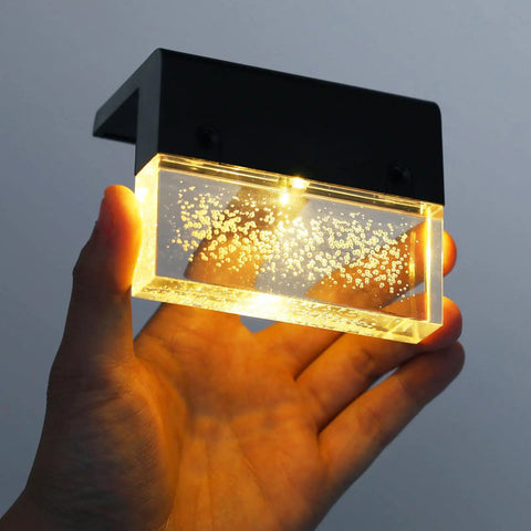 Hand holding a 3000K warm white color changing LED fixture, part of Solar Deck Lights 3000K Warm White Color Changing Outdoor Fence Lights For Steps Rails Walls Poolside LED Waterproof Patio Decor 6 Pack No Fuss Install Durable Weatherproof Solar Power, illuminated to show bubbles in the glass.