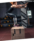 Person performing a box jump in gym with visible kettlebells and medicine balls in the background