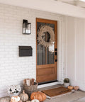 Outdoor Black Wall Sconce Light Fixture With Sturdy Iron Frame And Clear Glass Shade next to wooden front door with autumn decor