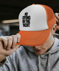 Man wearing orange and white trucker hat with 'No Crying on the Yacht' text, adjusting brim, promoting casual outdoor style.
