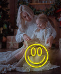Happy Face Neon Sign LED Neon Light Wall Decor USB Powered Yellow Neon for Bedroom Kids Room Wedding Party Brighten Your Space With A Fun Modern Accent And Everyday Vibe placed near a mother and child opening gifts in a cozy room.