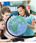 Children examining a globe on a stand in a classroom setting with bookshelves in the background.