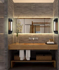 Modern bathroom with wooden vanity, brick wall tiles, and wall-mounted lights beside a mirror, accented with plants and towels.