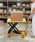 Solid Frame Hydraulic Scissor Lift Table Cart With Five Hundred Pound Capacity And Four Quiet Rollers raising a box in a warehouse.