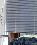 Interior view of window with closed white blinds and a plastic pull cord against a blue sky backdrop.