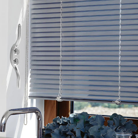 Interior view of window with closed white blinds and a plastic pull cord against a blue sky backdrop.