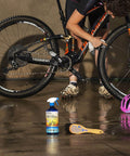 Person cleaning bike with Trail Ready Eco Friendly Bike Care Bundle, bottles of wet lube, degreaser, and cleaner visible on floor.