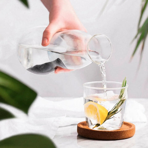 Hand pouring water from carafe with Charcoal Water Filter Sticks For Dope Tasting Hydration Inspired By Old Japanese Tradition Lasts Six Months Per Stick Perfect For Your Reusable Water Bottle And Everyday Sips into glass with lemon slice