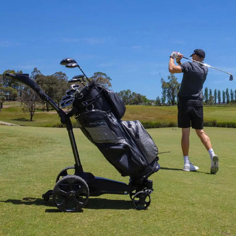 Slick Remote Controlled Electric Golf Push Cart Bundle With Top Shelf Touchscreen Gps Navigation And Preloaded Courses For Thirty Six Holes With Built In Drink Holder And Compact Umbrella Mount Plus Badass Travel Carry Bag next to golfer swinging club.