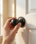 Hand holding a black cast iron doorknob on a white door, illustrating Two Pack Black Cast Iron Barn Door Handles With Vintage Distressed Texture Tough As Nails Heavy Duty Round Pulls For Shed Doors And Garage Rustic Industrial Style Hardware For Garden And Home Use.