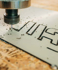 Close-up of a spiral router bit cutting a white panel with carved HARF letters, wood shavings around.