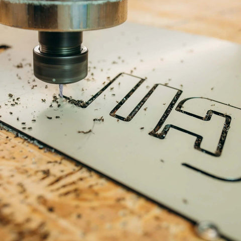 Close-up of a spiral router bit cutting a white panel with carved HARF letters, wood shavings around.