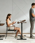 Solid Mobile Standing Desk With Pneumatic Lift And Tilt Top, Smooth Swivel Casters, Heavy Duty Steel Frame Walnut Laptop Workstation For Home Office Flex Bedside Chill used by a woman reading and a man working.