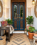 Charming entryway with woven rug, potted plants, and decorative mirrors flanking a wooden bench and ornate blue double doors.