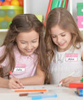 Two girls wearing Light Pink Hello My Name Is Stickers Set, 300 Pcs Name Tag Labels On A Roll For Back To School drawing together.