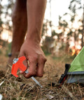Person installing Rock Solid Heavy Duty Metal Tent Stakes Set with 7.87 Inch Screw In Pegs and bright orange plastic heads near tent on grass.