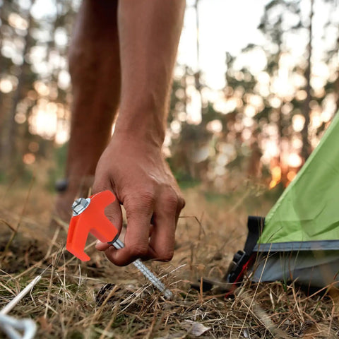 Person installing Rock Solid Heavy Duty Metal Tent Stakes Set with 7.87 Inch Screw In Pegs and bright orange plastic heads near tent on grass.