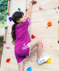 Child climbing wooden wall with colorful handholds, dressed in purple matching top and shorts, wearing white sneakers.