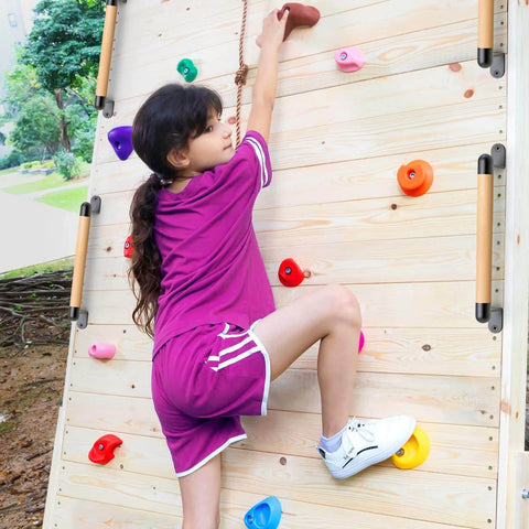 Child climbing wooden wall with colorful handholds, dressed in purple matching top and shorts, wearing white sneakers.