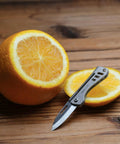 Titanium Mini Pocket Knife With Bottle Opener For EDC And Outdoor Gear next to sliced orange on wooden surface