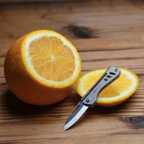 Titanium Mini Pocket Knife With Bottle Opener For EDC And Outdoor Gear next to sliced orange on wooden surface