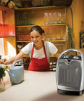 Oscillating Ceramic Tabletop Space Heater With Adjustable Thermostat Carrying Handle Two Quiet Heat Settings And Fan Only Mode 11.6 Inches Silver Compact Space Saving Home Heating Solution on a kitchen countertop with a woman smiling in the background.