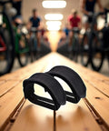 2 Pack Black Pedal Straps for Kids Bikes and Fixed Gear on a wooden surface with cyclists in the background, durable toe clip straps.