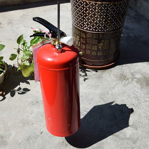 Red fire extinguisher outdoors beside potted plants and decorative container on concrete surface.