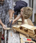 Child using saw to cut wood with assistance, wooden toy car in foreground, tools scattered on workshop table.