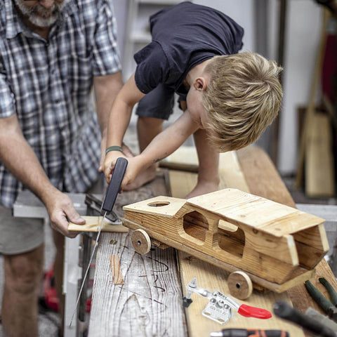 Child using saw to cut wood with assistance, wooden toy car in foreground, tools scattered on workshop table.