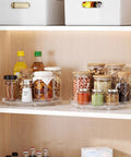 Nine Inch Clear Lazy Susan Set Of Two Round Rotating Turntable Organizers With Non Slip Base For Pantry Cabinet And Fridge Shelf organizing spices and snacks in jars on a shelf.