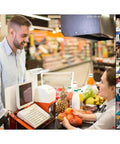 A cashier interacting with a customer at a grocery store checkout with fruits and beverages visible.