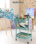 3 Tier Rolling Cart With Lockable Wheels And Hanging Cups in studio setting with art supplies, green metal, airflow mesh baskets