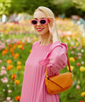 Woman in pink dress and sunglasses holding an orange bag, standing in a colorful flower garden