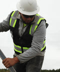 Construction worker wearing a Ridgeline Full Brim Hard Hat With 4 Point Ratchet Suspension, grey jacket, safety vest, holding a tool on site.