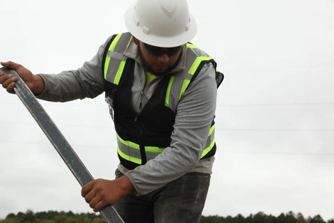 Construction worker wearing a Ridgeline Full Brim Hard Hat With 4 Point Ratchet Suspension, grey jacket, safety vest, holding a tool on site.