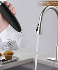 Handheld Electric Milk Frother Wand for Coffee at Home being rinsed under a kitchen faucet, stainless steel whisk visible, black handle.