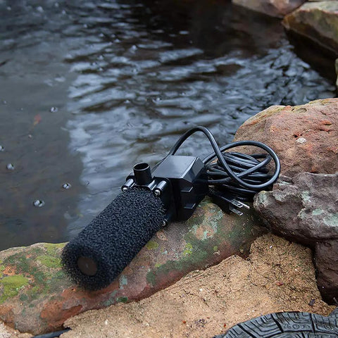 Black FR900 Pond Pump Set Submersible Fountain Pump For Outdoor Water Features, positioned on rocks by a pond edge
